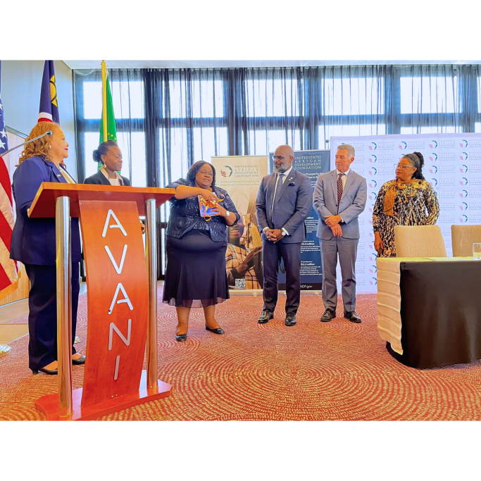 Five people stand at a podium during a formal event with flags in the background.